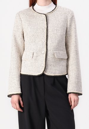 Frau trägt eine cremefarbene Tweed-Cropped-Jacke mit schwarzem Besatz, Knopfverschluss und zwei vorderen Klappentaschen über einem weißen Top und schwarzen Hosen.