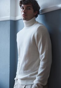 Bela turtleneck pulover z mehkimi teksturami, z rebrastim ovratnikom in manšetami, v kombinaciji s svetlimi hlačami pred modro steno.