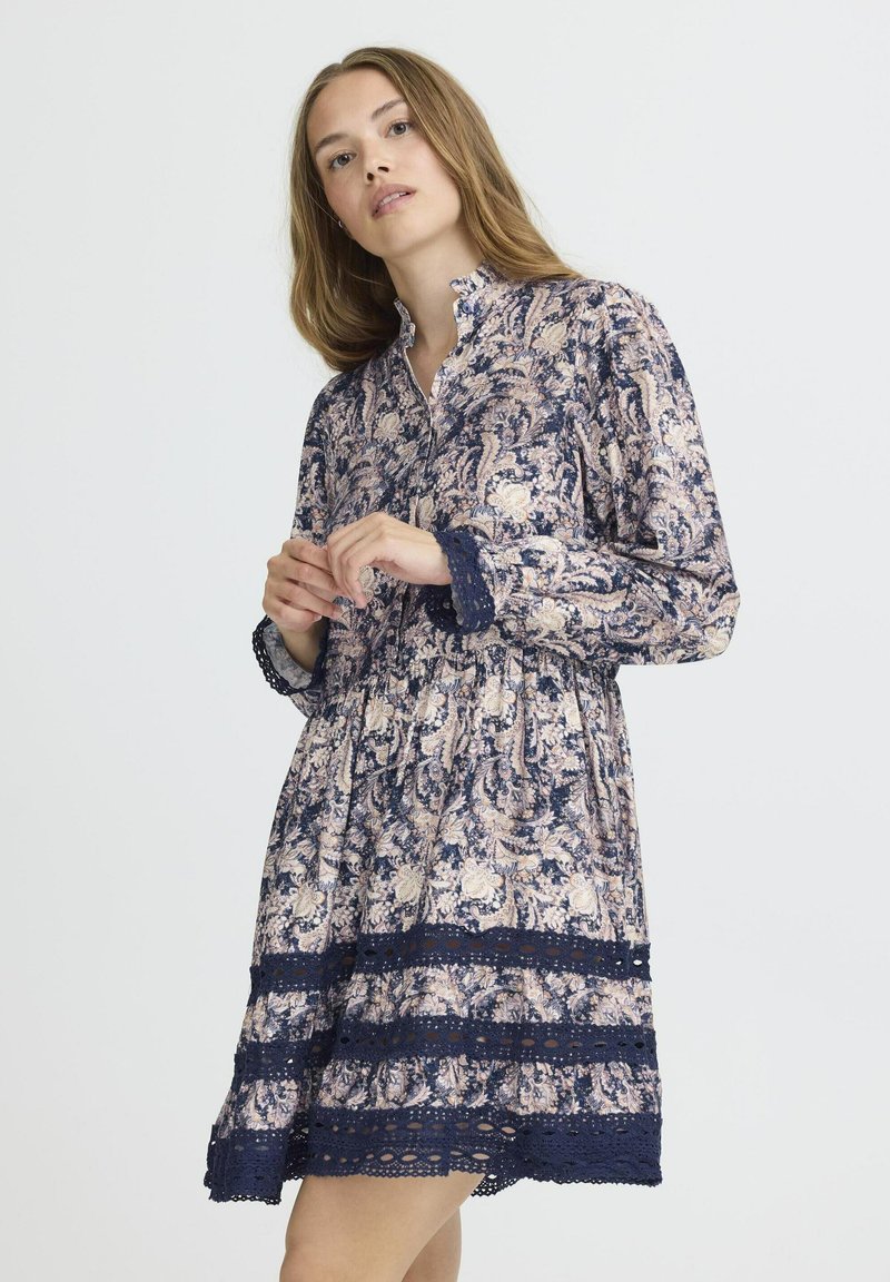 Garais piedurknes kleita jūras zila krāsā ar rozā un krēmkrāsas ziedu rakstu. Tam ir apkakles izgrieza un mežģīņu akcenti apakšmalā un piedurkņu galos.