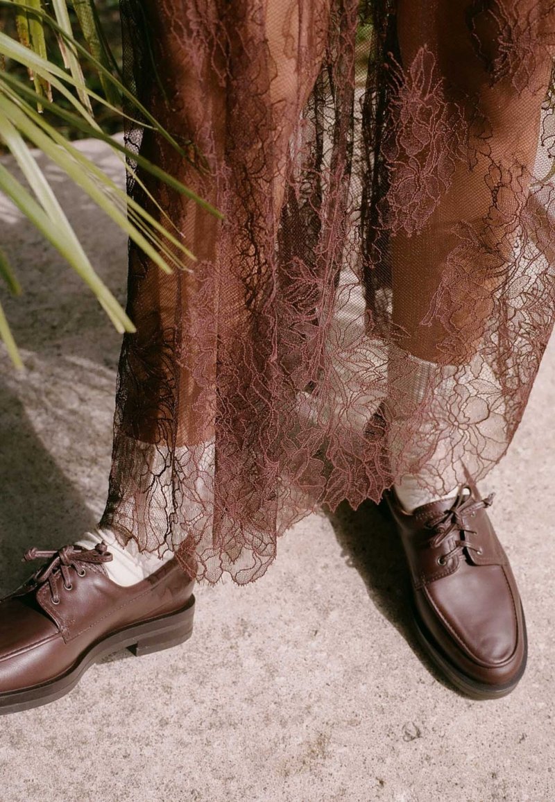 Jambes en chaussettes blanches et chaussures en cuir marron à lacets sous une jupe en dentelle brune transparente, debout sur du béton près de feuilles de plantes vertes.