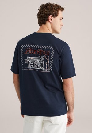 Marineblauw katoenen T-shirt met een grafisch ontwerp van een winkelpand met de tekst "BLUE RIDGE" in oranje, omgeven door een dambordpatroon.