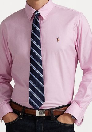 Mand iført en pink skjorte med knapper, brun bælte, blå stribet slips og mørke jeans med hænderne i lommerne.