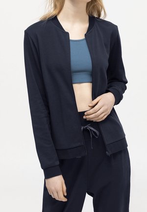 Femme portant une veste zippée bleu marine sur une brassière de sport bleue et un pantalon bleu marine à cordon, debout devant un fond uni clair.