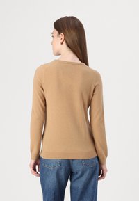 Svetlo rjav pulover, izdelan iz mehke, teksturirane tkanine. Ima dolge rokave in okroglo ovratnico, v kombinaciji z modrimi jeans hlačami.