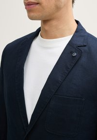 Blazer din in marin cu un singur buzunar și detaliu cu nasture, purtat peste un tricou alb cu guler rotund. Prim-plan care arată gulerul și textura materialului.