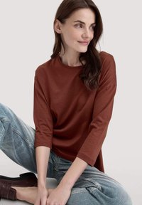Frau mit langen, dunklen Haaren, die ein rostfarbendes Langarmshirt und hellblaue Jeans trägt, im Schneidersitz sitzt, zur Seite schaut und leicht lächelt.
