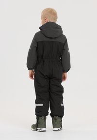 Salopette extérieure noire avec des accents gris, taille élastique et bandes réfléchissantes. L'enfant porte des bottes vert olive. L'arrière-plan est blanc uni.