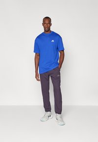 Blue cotton t-shirt with white logo, paired with dark gray pants and white sneakers with blue accents. Plain background.