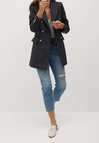 Kostkovaný blazer v tmavě šedé barvě se stříbrnými knoflíky, zkombinovaný s různě opotřebovanými modrými džíny a bílými slip-on botami.
