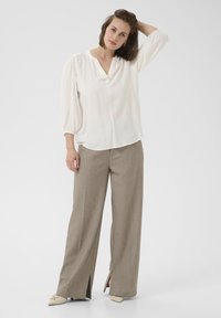 Blusa blanca con mangas abullonadas y escote en V, combinada con pantalones de pierna ancha color marrón claro con aberturas laterales. Patrón texturizado sutil.