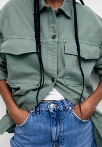 Light green, button-up shirt with two chest pockets and rolled sleeves, paired with denim shorts featuring a high waist and frayed hem.