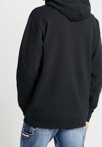 Persona viste una sudadera negra con capucha sencilla combinada con jeans de mezclilla claros, vista desde atrás sobre un fondo liso.