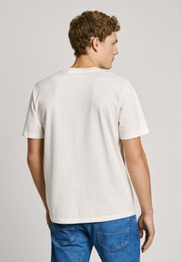 Short-sleeve, crew-neck t-shirt in off-white cotton with a smooth texture, displayed from the back, paired with blue denim jeans.