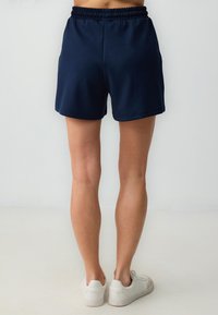 Een persoon die staat, draagt een marineblauwe korte broek en witte sneakers, met de benen zichtbaar vanaf de taille naar beneden tegen een neutrale achtergrond.