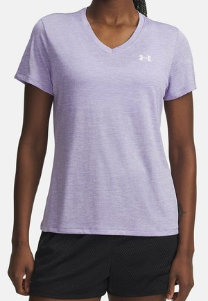 Kobieta nosząca jasnofioletowy T-shirt z krótkim rękawem i dekoltem w serek z białym logo Under Armour na piersi oraz czarne szorty z siateczki.