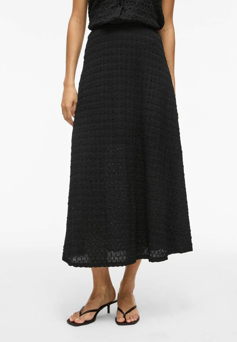 Femme portant une jupe midi noire texturée avec un top assorti et des sandales noires à talons, debout devant un fond blanc uni.