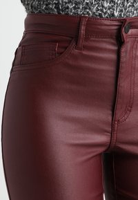 Pantalones ajustados de piel sintética burdeos, con bolsillos delanteros y costuras tono sobre tono, destacando una textura suave y brillante.