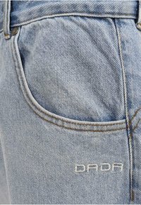 Šviesiai mėlyni džinsai su matomu siuvimu, priekinė kišenė ir išsiuvinėtas "DADA" logotipas. Medžiaga turi tekstūruotą audimo raštą.