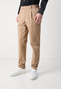 Béžové lehké chino kalhoty s rovným střihem, vybavené bočními kapsami a plochým předním dílem, zkombinované s bílými teniskami.