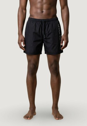 Torso e gambe maschili indossano pantaloncini da bagno neri con la scritta "Armani Exchange" stampata verticalmente sul lato destro, in piedi a piedi nudi su uno sfondo neutro.