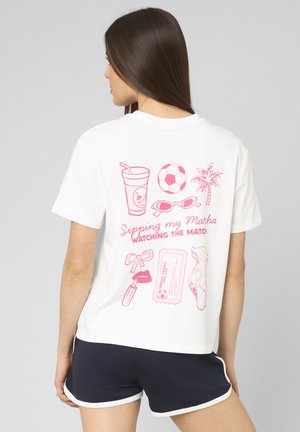 Femme portant un t-shirt blanc avec des motifs roses : boisson, ballon de football, palmier, lunettes de soleil, rouge à lèvres, billet de match et sandales, assorti à un short bleu marine.