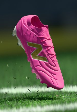 Leuchtend pinker Fußballschuh mit goldenem "N"-Logo, der auf einem markierten grünen Spielfeld Gras aufwirbelt.