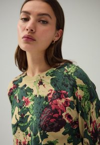 Beige pullover met bloemenpatroon en groene en rode accenten. Heeft een geribbelde halslijn en een zachte textuur. Casual pasvorm met lange mouwen.