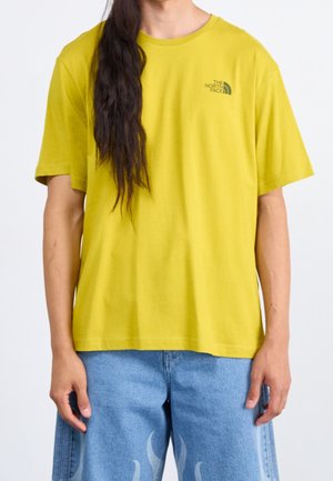 Person iført gul The North Face t-shirt og lyseblå jeans med flammedesign, står med afslappede arme langs siden.