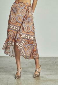 Jupe asymétrique marron et bleue à motifs avec un ourlet volé et des designs géométriques. Portée avec des talons métalliques argentés à brides croisées.