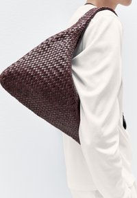 Vävd vinröd axelväska med triangulär form och texturerad yta, som har en enkel rem och minimalistiska designelement.