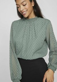 Kobieta uśmiechająca się, w zielonej bluzce z długim rękawem o zygzakowatej fakturze i zebranym wysokim dekoltem, w zestawie z czarnymi spodniami.