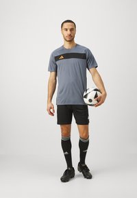 Szary sportowy T-shirt z czarnym paskiem i pomarańczowym akcentem, połączony z czarnymi szortami, czarnymi skarpetkami w białe paski i czarnymi korkami, trzymający piłkę nożną.