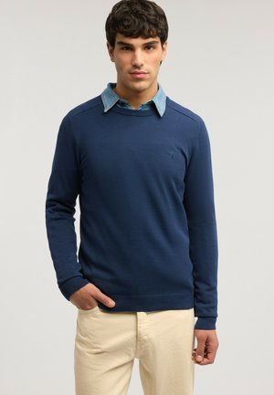 Joven vestido con un suéter azul marino sobre una camisa de cuello azul claro y pantalones beige, de pie frente a un fondo claro y liso.