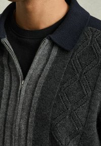 Svetr na zip v tmavě šedé a černé vlněné směsi, se vzorovaným texturou a žebrovaným tmavě modrým límcem. Obsahuje stříbrný detail zipu.