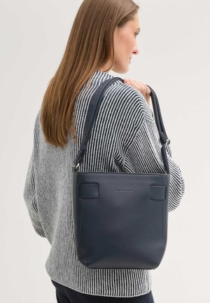 Femme aux longs cheveux bruns portant un pull à rayures, tenant un sac à bandoulière en cuir bleu marine sur son épaule droite, sur un fond clair.