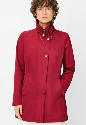 Frau trägt eine rot Jacke mit langen Ärmeln und hohem Kragen mit Druckknöpfen, eine Hand in der Tasche, vor einem weißen Hintergrund stehend.