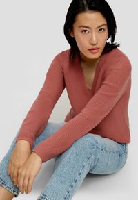 Roodbruin gebreid sweater met een V-hals en ribbels, gecombineerd met lichtblauwe denim jeans, zittende houding, handen op de knieën gevouwen.