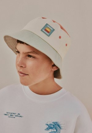 Berniukas vilki kreminės spalvos kibiro formos skrybėlę su saulės ženkleliu ir baltą džemperį su mėlynu japonišku užrašu bei drakono piešiniu.