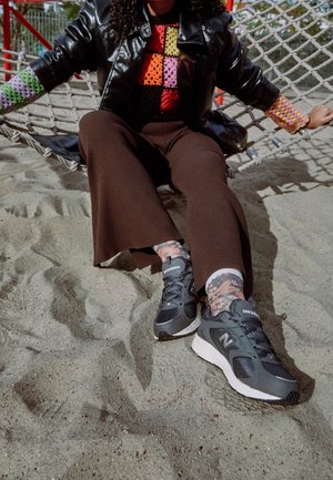 Person sitzt in einer Netz-Hängematte und trägt einen bunten Patchwork-Pullover, dunkelbraune Hose, florale Socken und schwarze New Balance Sneakers auf sandigem Boden.