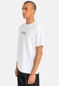 White cotton t-shirt with short sleeves, round neckline, and a black patterned graphic design on the chest, paired with dark jeans.