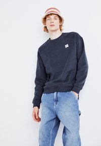 Navy blue sweatshirt with crew neckline and ribbed cuffs, featuring a small white logo. Worn with light blue baggy jeans.