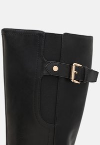 Bota de cuero negro hasta la rodilla con un acabado suave, paneles laterales elásticos y un accesorio de hebilla en tono dorado en la correa.
