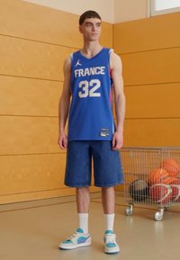 Maglia sportiva blu con "FRANCIA" e numero "32," abbinata a pantaloni shorts in denim e sneakers blu con dettagli bianchi e viola.