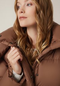 Brązowa puchowa kurtka z wysokim kołnierzem, wykonana z gładkiego materiału. Zawiera zamek błyskawiczny oraz zapięcie na zatrzask, a pod spodem znajduje się strukturalny szary sweter.