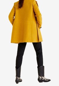 Geel wollen jas met een losse pasvorm, zwarte broek en zwarte cowboylaarzen met witte decoratieve accenten en textuurpatronen aan de bovenkant.