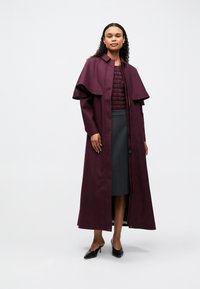 Sieviete ar gariem cirtainiem matiem, ģērbta plūmju krāsas garā mētelī pār atbilstošu topu un pelēkiem svārkiem, stāv uz neitrālas gaismas fona.