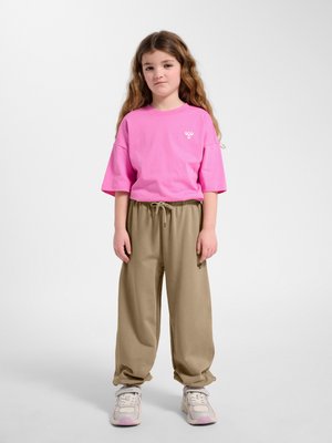 Ung pige står med afslappet holdning iført lyserød oversized t-shirt, beige brede bukser og hvide sneakers på ensartet hvid baggrund.