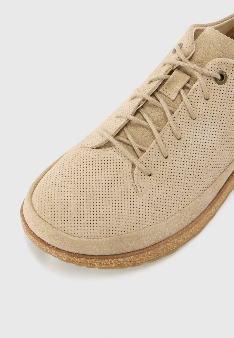 Helle beige Wildlederschuhe mit perforiertem Obermaterial, flachen Schnürsenkeln und einer strukturierten braunen Sohle. Abgerundete Zehenform und minimalistisches Design.