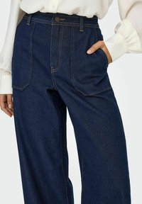 Un pantalon en denim bleu foncé avec une coupe décontractée, comportant des poches avant et arrière et des coutures contrastées, présenté avec un haut neutre.