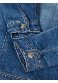 Detail džínové bundy s modrou látkou, kontrastním žlutým šitím a stříbrnými knoflíky na rukávech. Hladká textura.